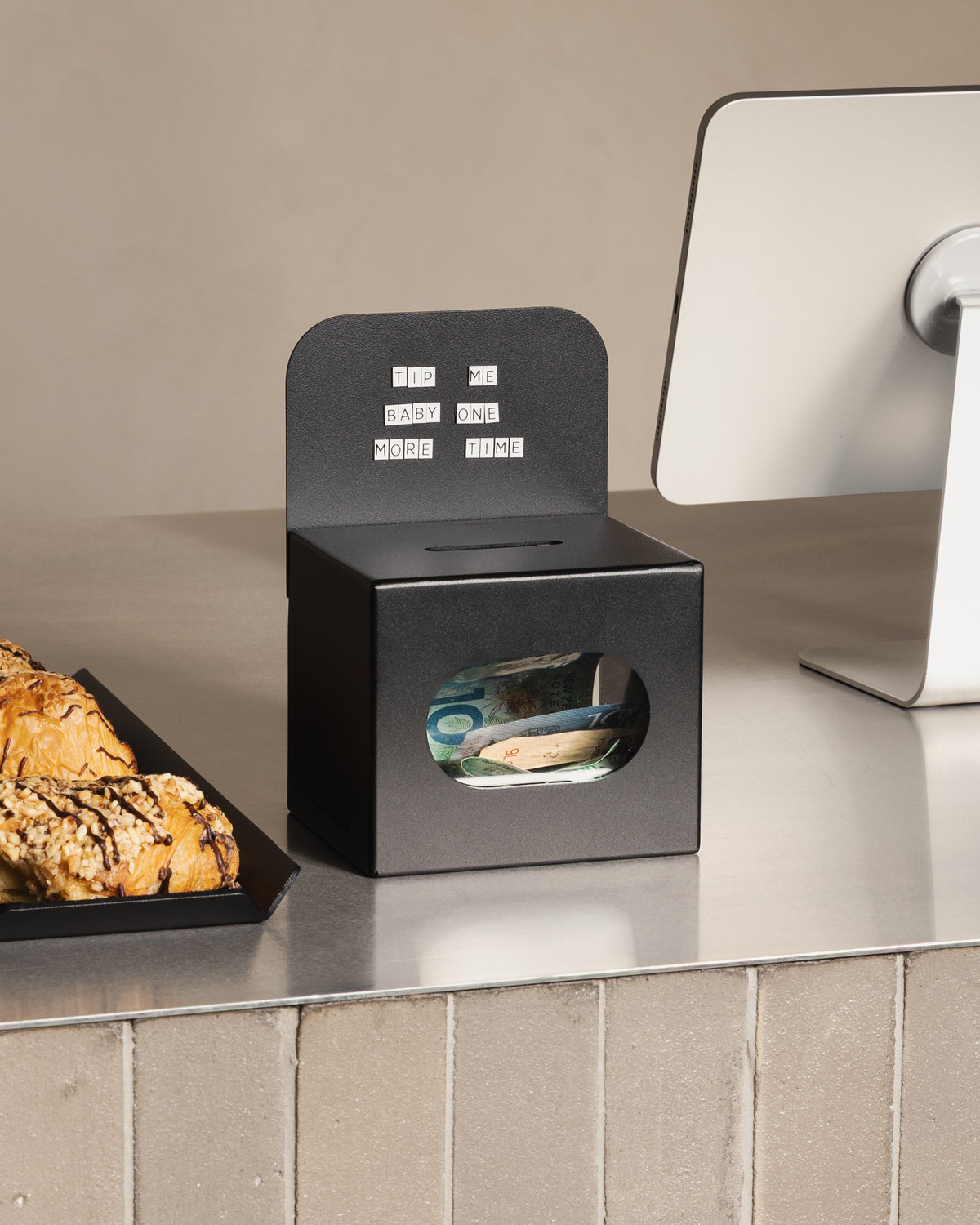 Black FOLD Tip Box by Made of Tomorrow on a café counter beside pastries and a payment terminal